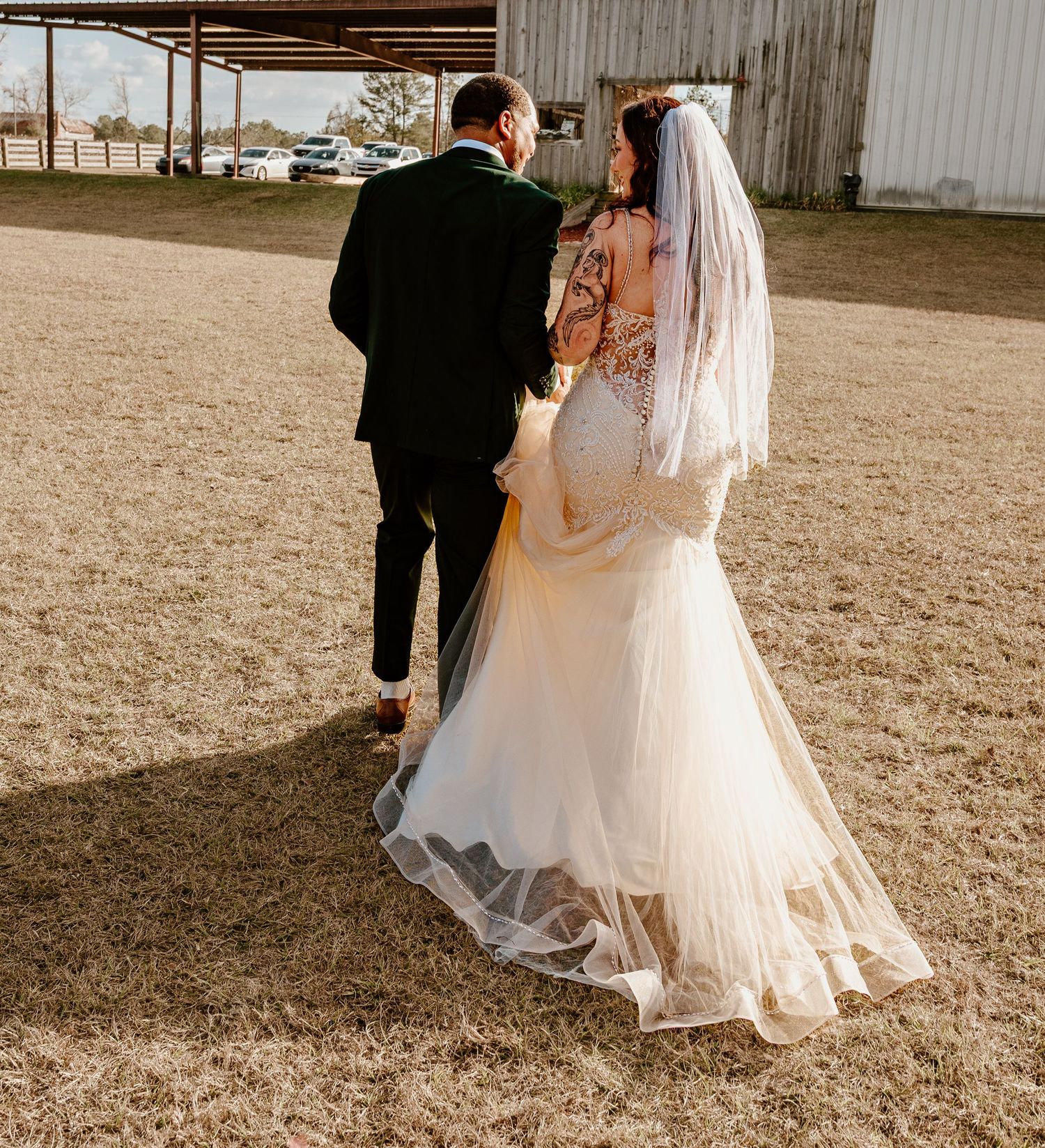 A bride and groom walking together in a rustic outdoor setting with a veil flowing in the breeze.