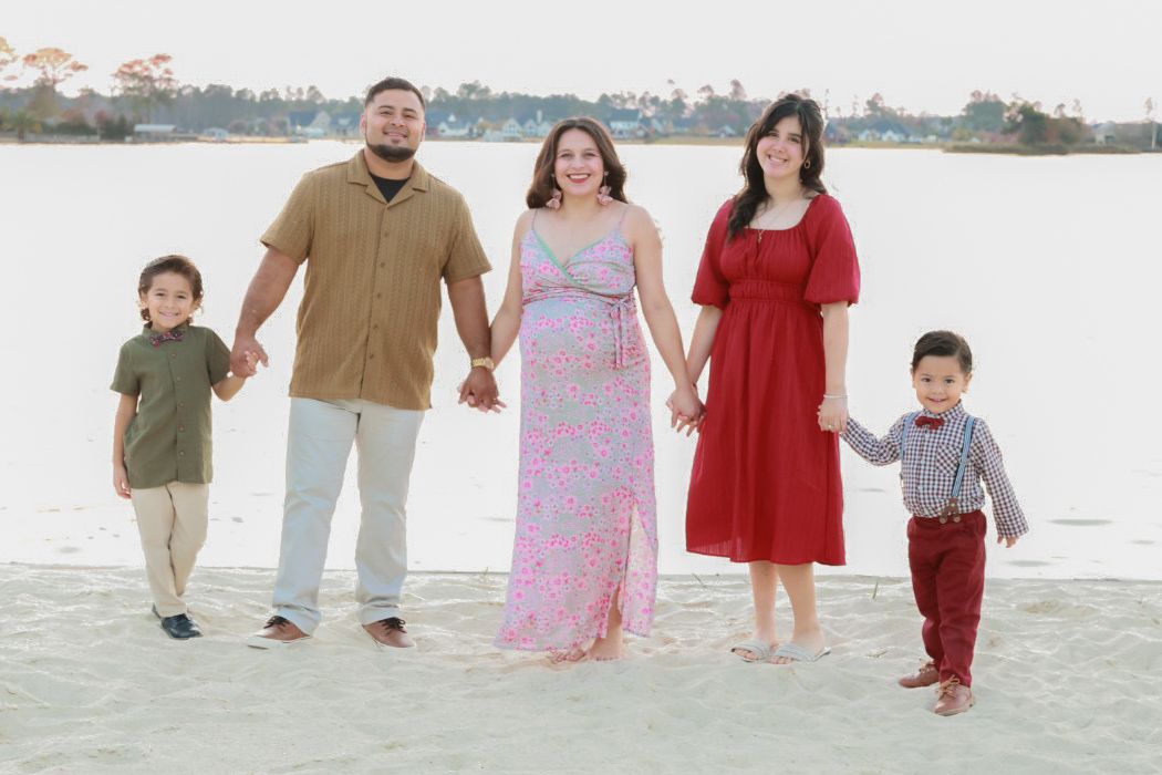A family wearing coordinated outfits poses together on a sandy beach at sunset with water in the background.