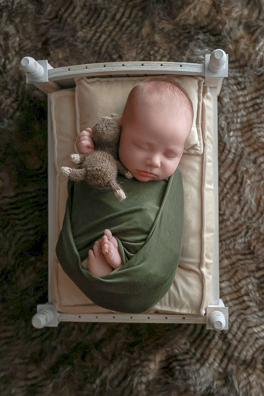 A sleeping newborn swaddled in green fabric rests in a white miniature bed frame with a small teddy bear companion.