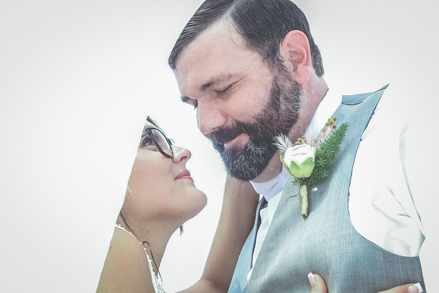 A bearded groom in a gray vest with boutonniere leans forehead-to-forehead with his bride wearing glasses on their wedding day.