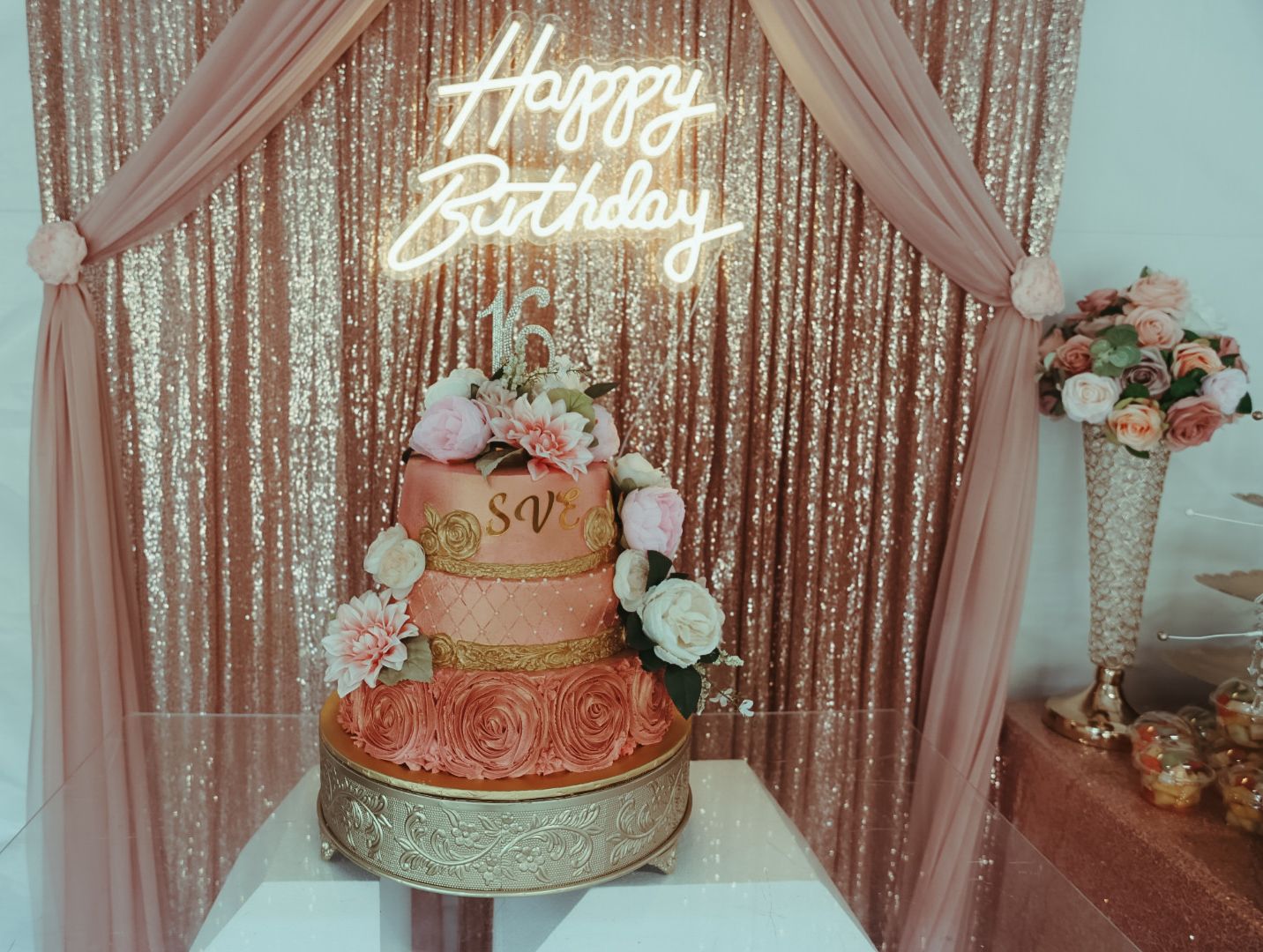 Three-tier pink and gold birthday cake with floral decorations under a Happy Birthday neon sign on a sequin backdrop.