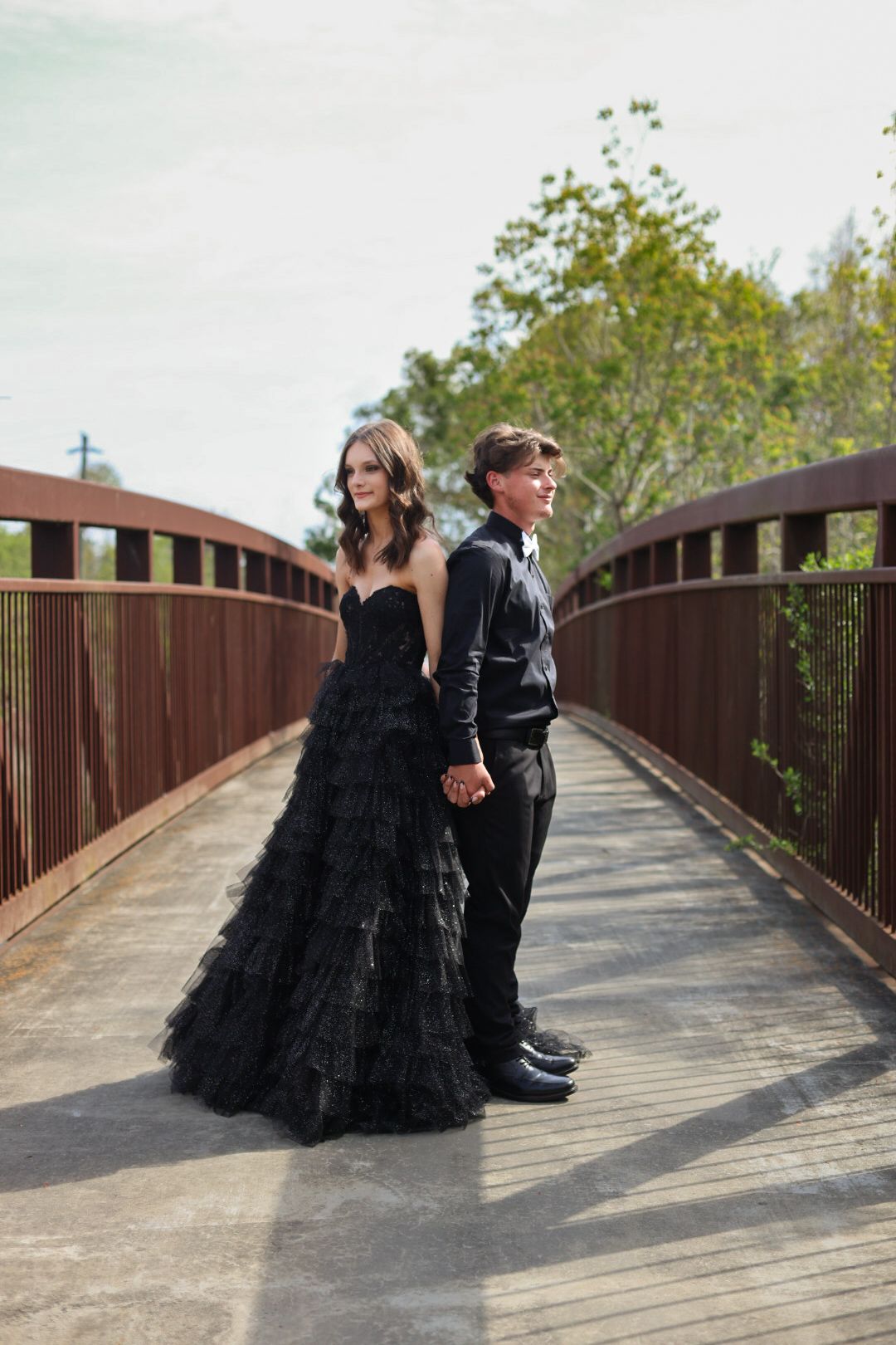 Couple in matching black formal wear holding hands on a wooden bridge surrounded by lush green trees.