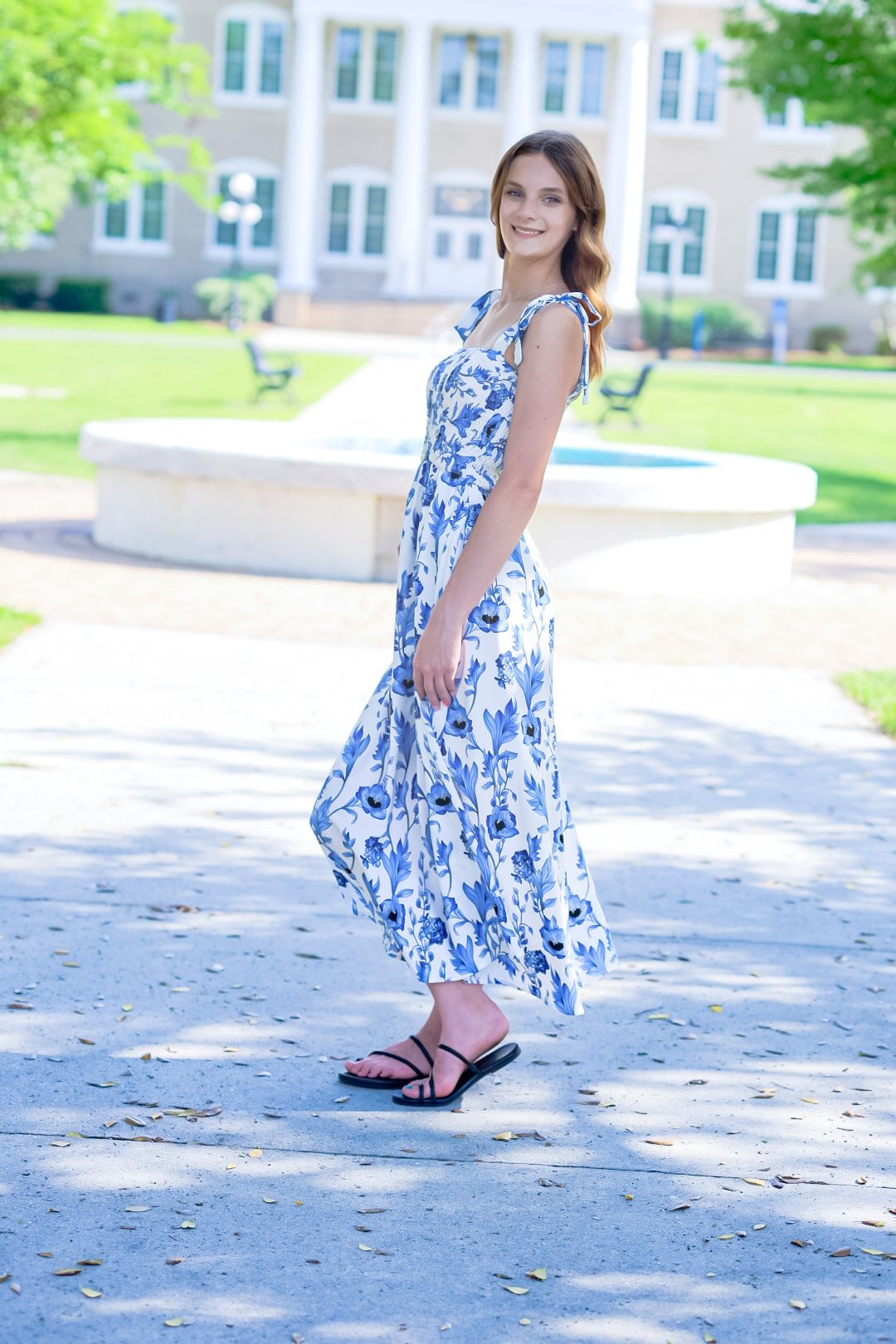 Smiling young woman in blue floral maxi dress twirling on sunny campus walkway.