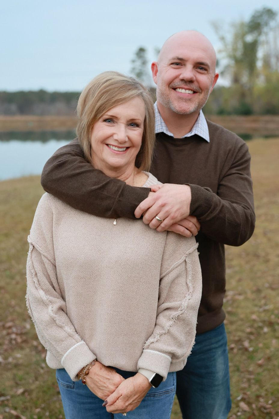 A couple embraces while standing outdoors near a lake on an autumn day, wearing casual sweaters and jeans.