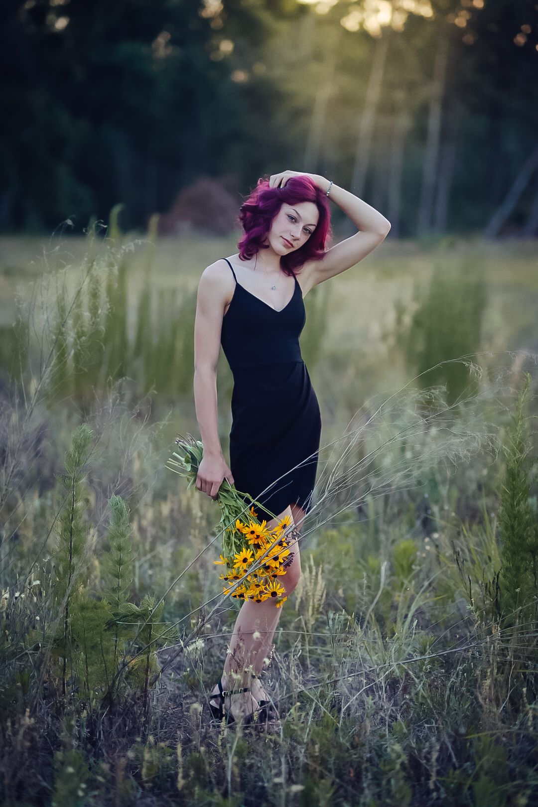A figure in a black dress stands in a sunlit meadow holding yellow wildflowers at sunset.