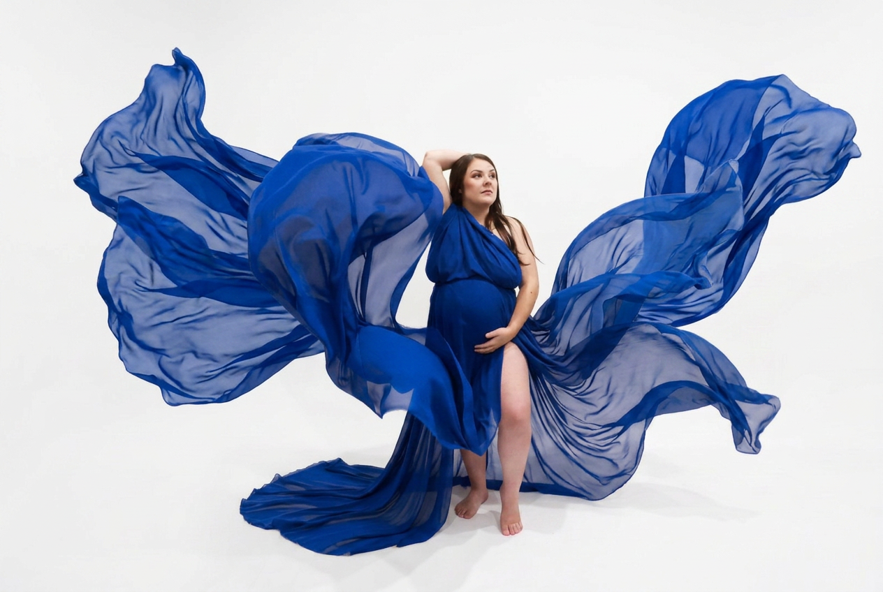 Pregnant woman in flowing royal blue chiffon gown with fabric billowing dramatically against white studio background.