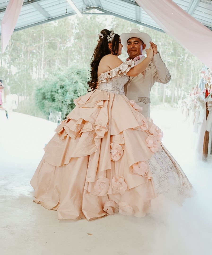 Couple dancing at a quinceañera, woman in a blush pink ball gown with floral details, man in a light gray suit.