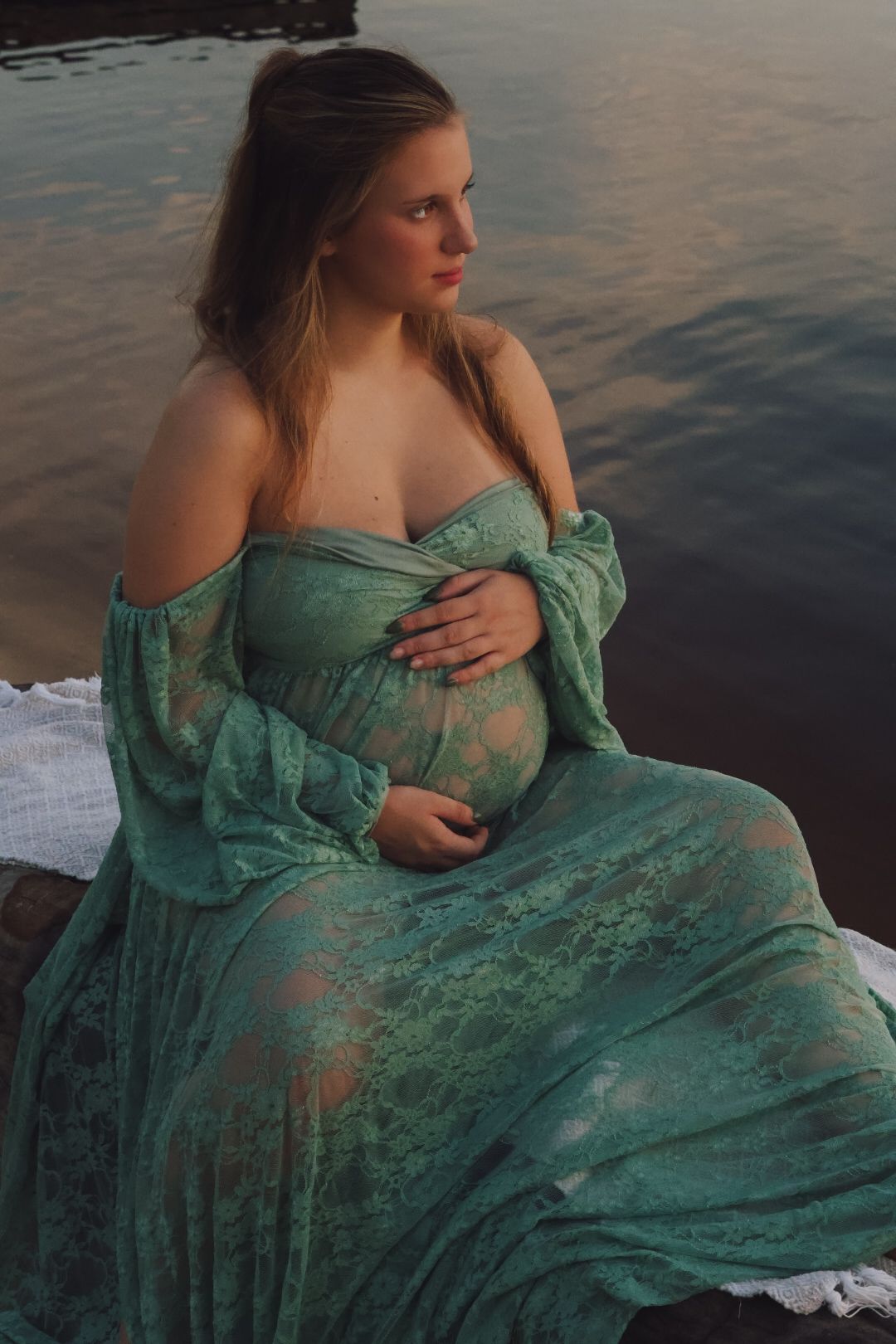 A serene maternity portrait by the water features a flowing seafoam green dress at sunset.