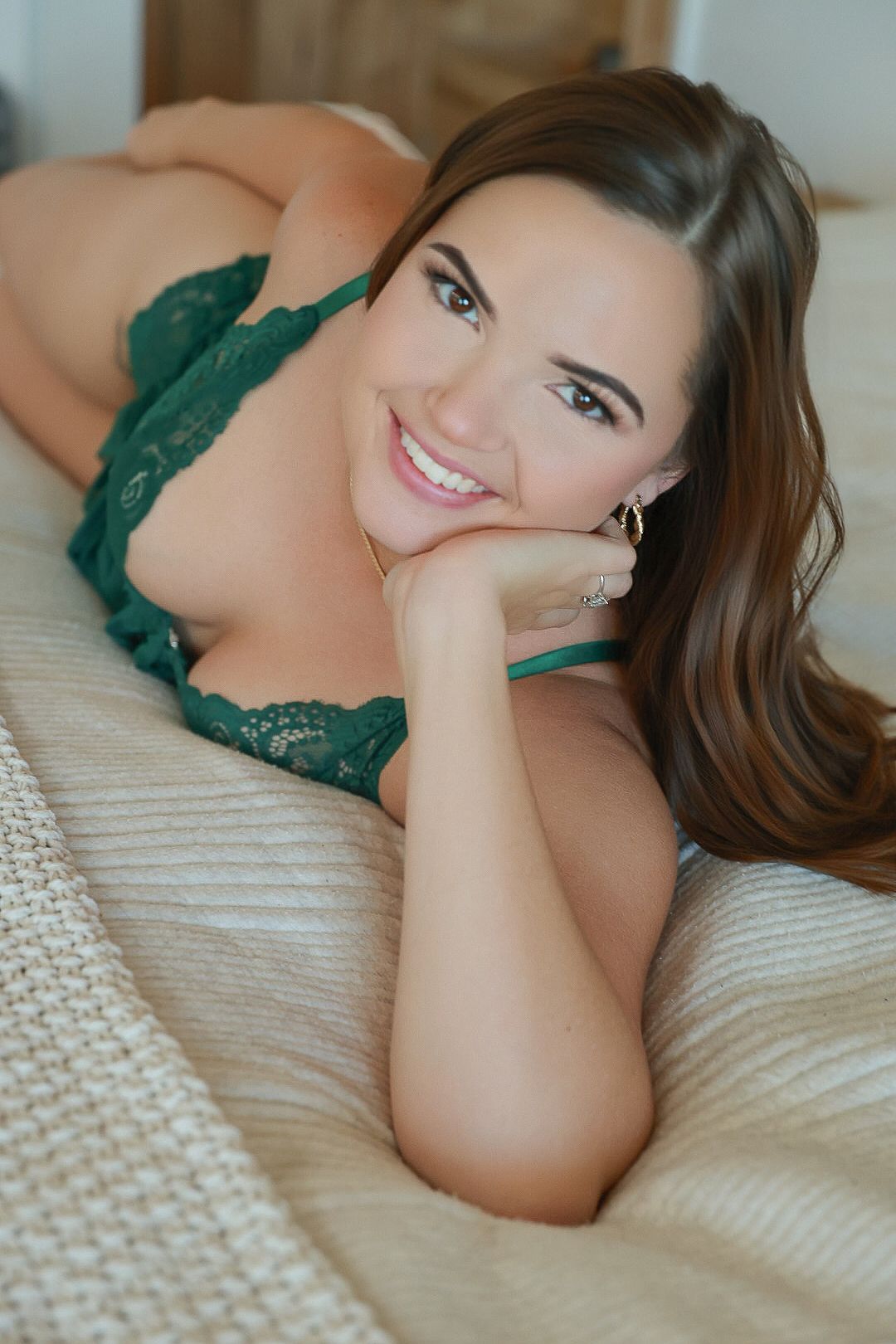 Smiling brunette woman in green lace lingerie lying on a bed, resting her chin on her hand.