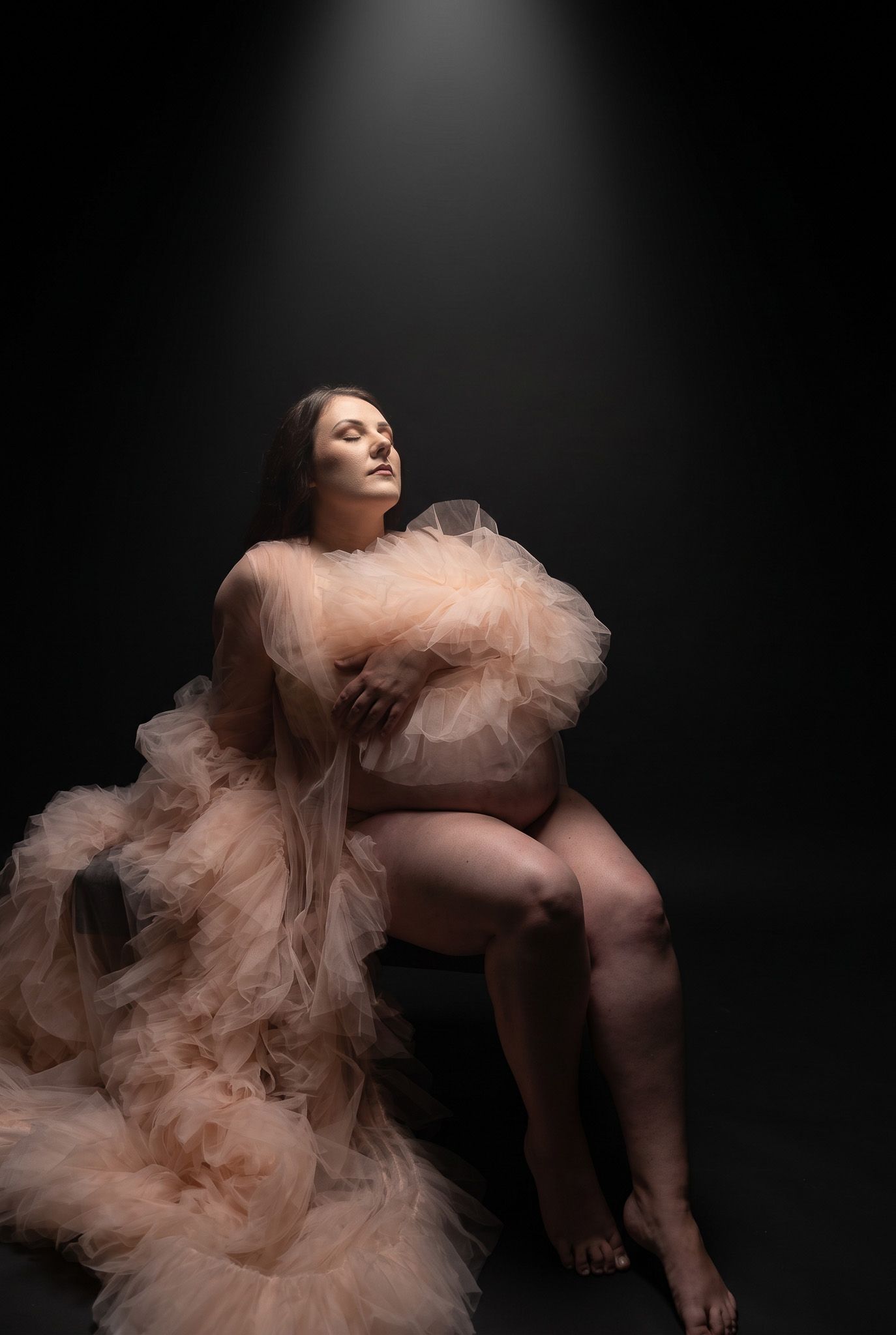 Pregnant woman in flowing peach tulle gown poses gracefully against dark studio backdrop in artistic maternity portrait.