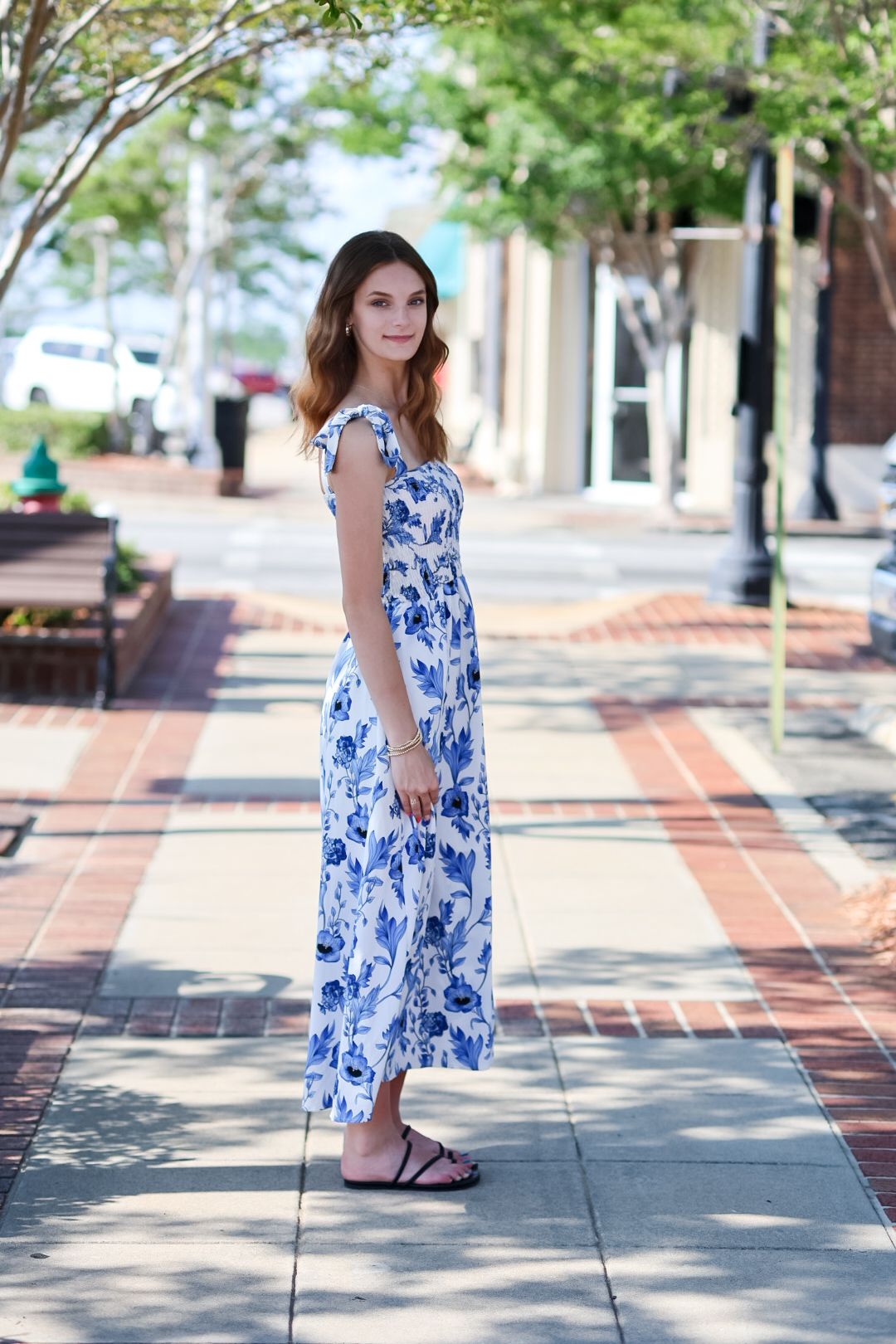 Young woman in white blue floral maxi dress standing on brick sidewalk in town.