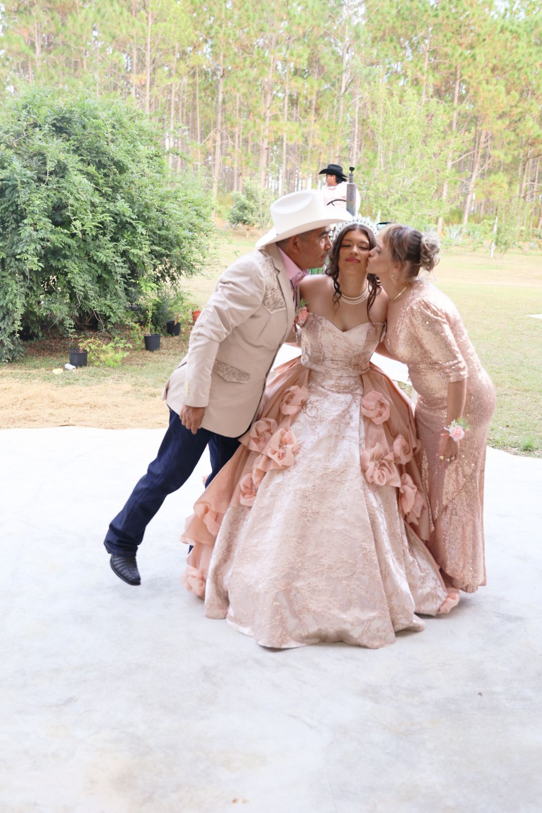 A young woman in a pink ball gown is kissed on the cheeks by two people at an outdoor celebration.