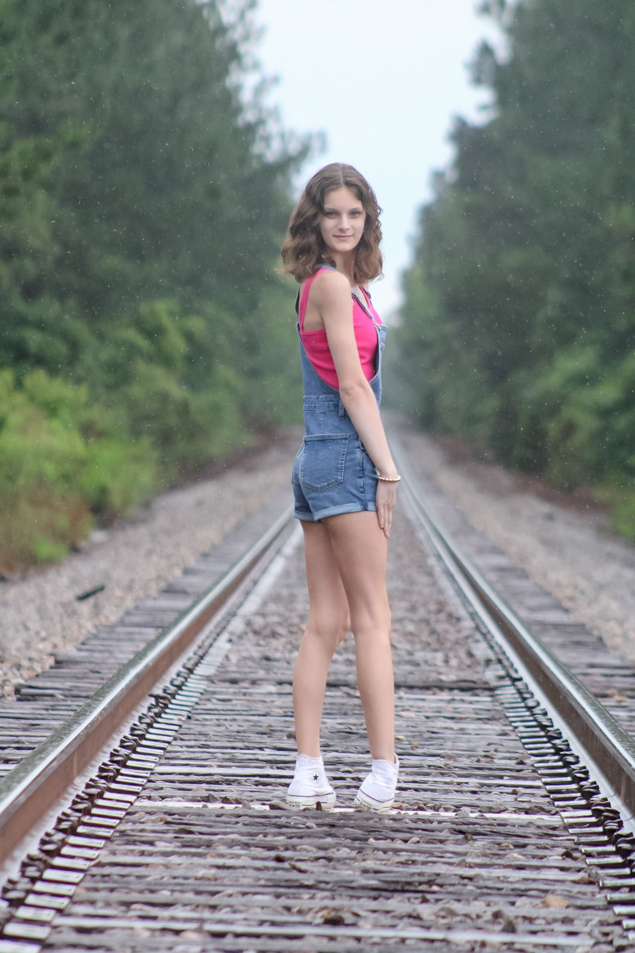 Young woman in denim overalls and pink top standing on railroad tracks surrounded by green trees.