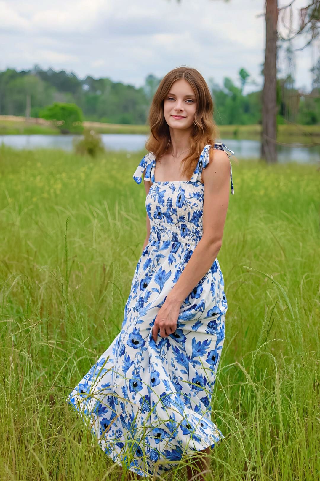 Young woman in blue floral maxi dress standing in tall green grass near a lake surrounded by trees.