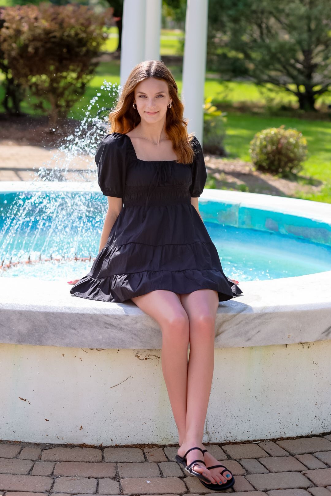 Young woman in black dress sitting on edge of outdoor fountain in a garden.