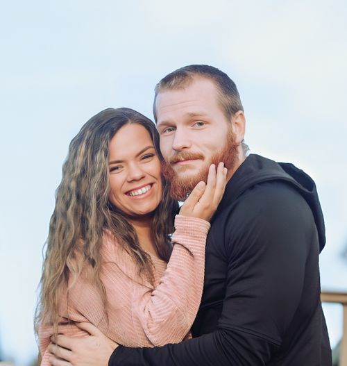 A smiling couple poses closely together outdoors, woman in pink sweater embracing man with red beard in black hoodie.