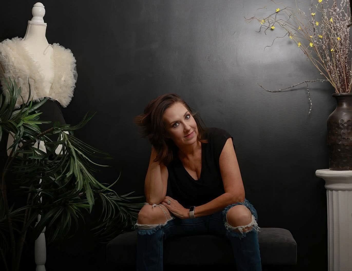 Woman in black top and ripped jeans sits pensively in a dark studio with plants and a dress form.
