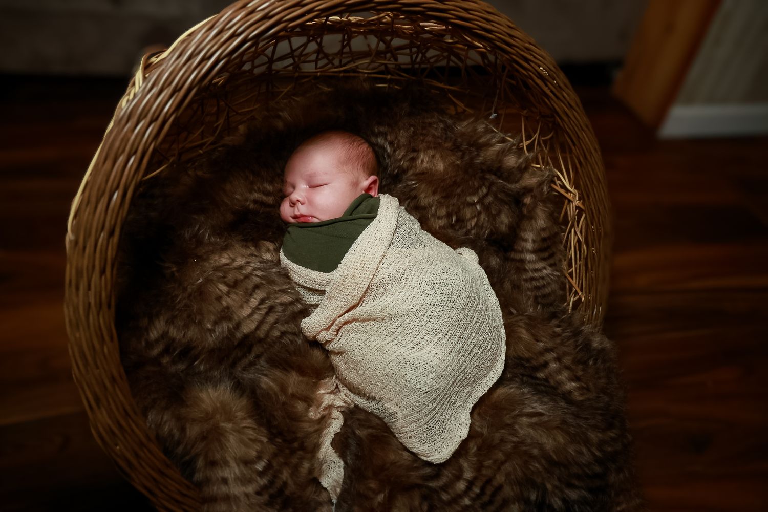 A newborn baby sleeps peacefully in a cozy brown wicker basket while wrapped in a soft cream-colored blanket.