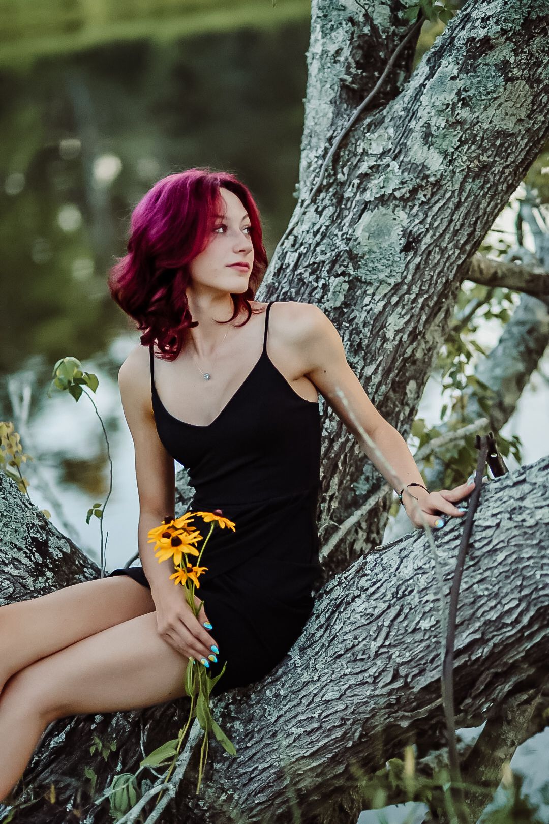 Figure with vibrant magenta hair poses on a tree branch holding yellow flowers in a natural outdoor setting.