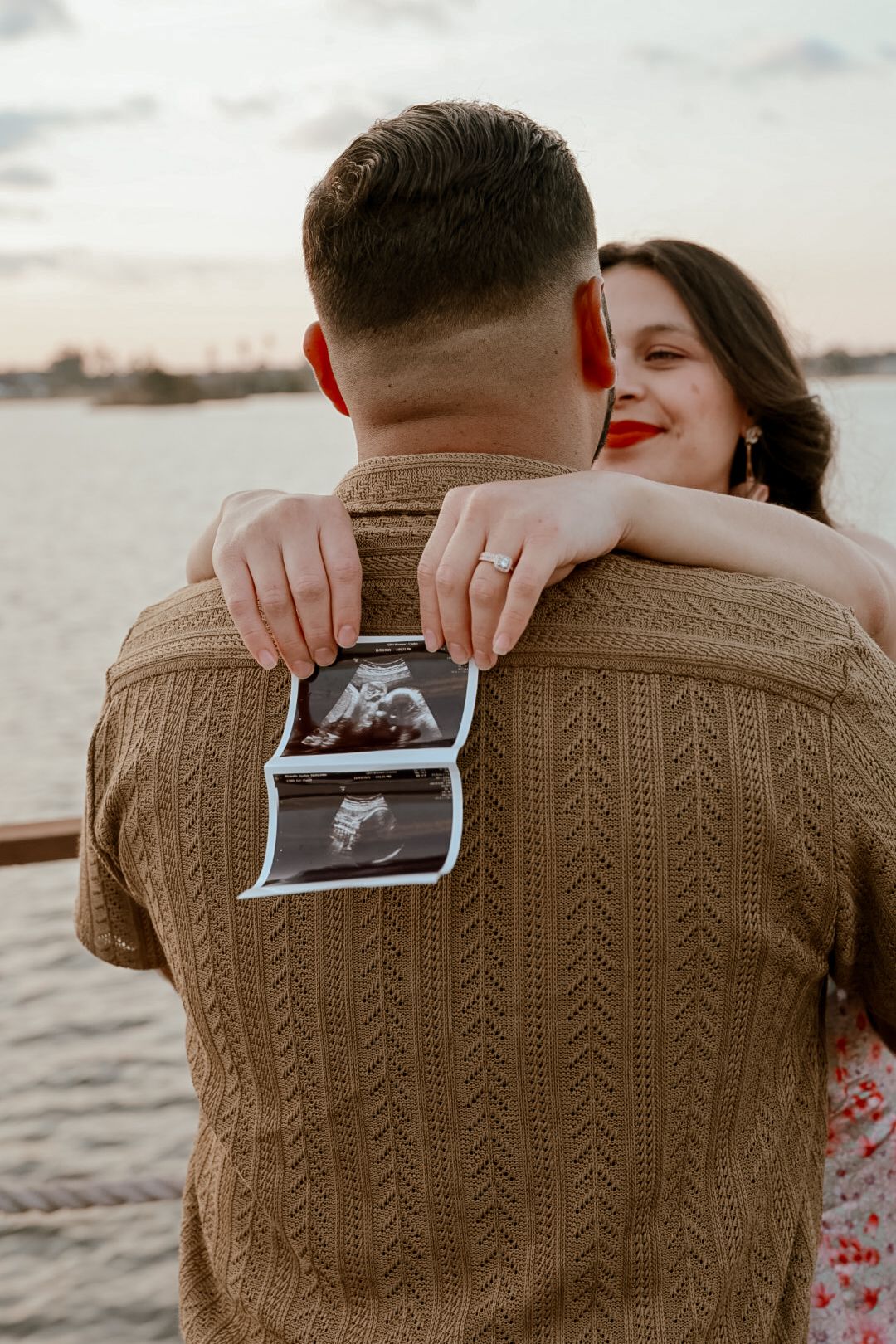 A couple shares a tender moment by the water while holding up an ultrasound photo against a brown knit sweater.