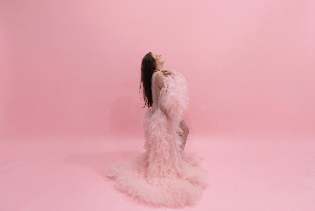 A woman poses in a stunning pink feathered gown against a soft pink studio background.