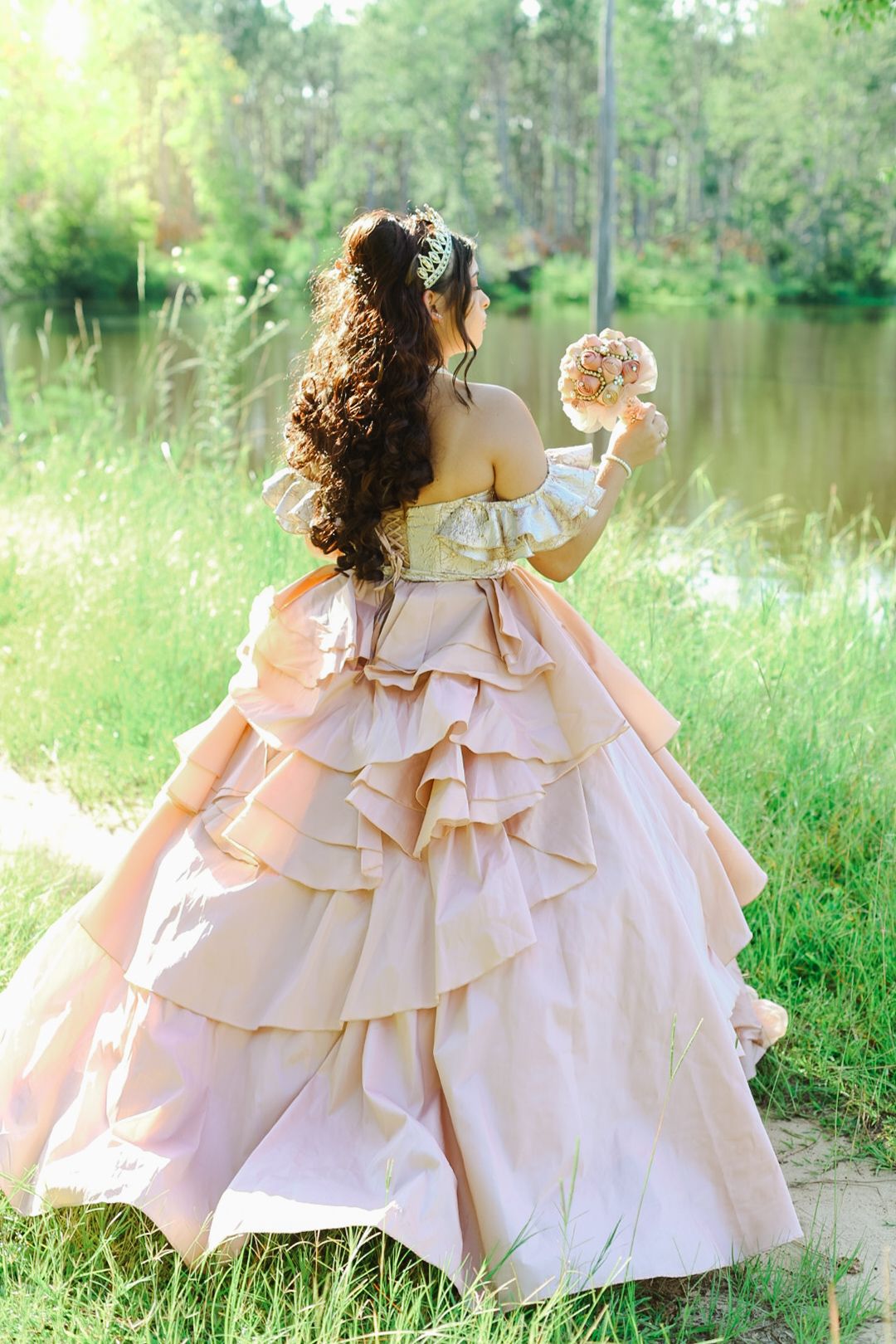 Woman in elegant pink ball gown with ruffled layers holds bouquet by serene lakeside surrounded by lush greenery.