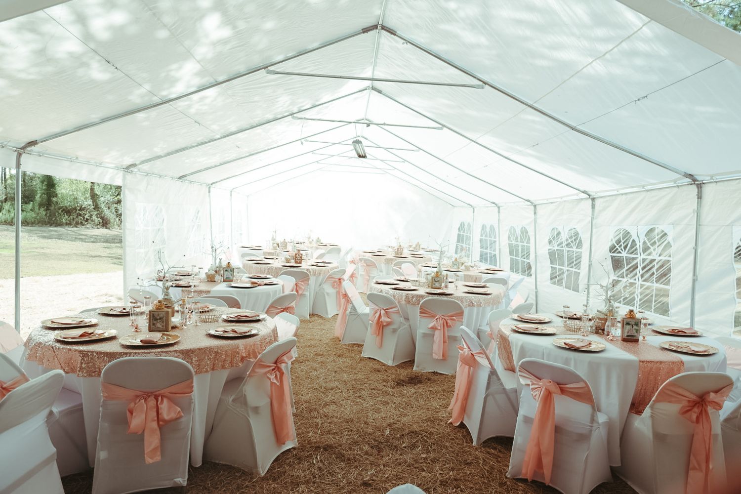 Elegant outdoor wedding tent reception with white chair covers, pink satin sashes, and decorated round tables on hay-covered ground.