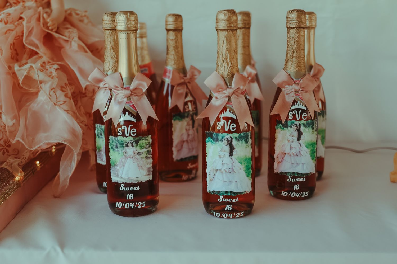 Four personalized Sweet 16 champagne bottles with pink ribbons and custom labels displayed on a white table.