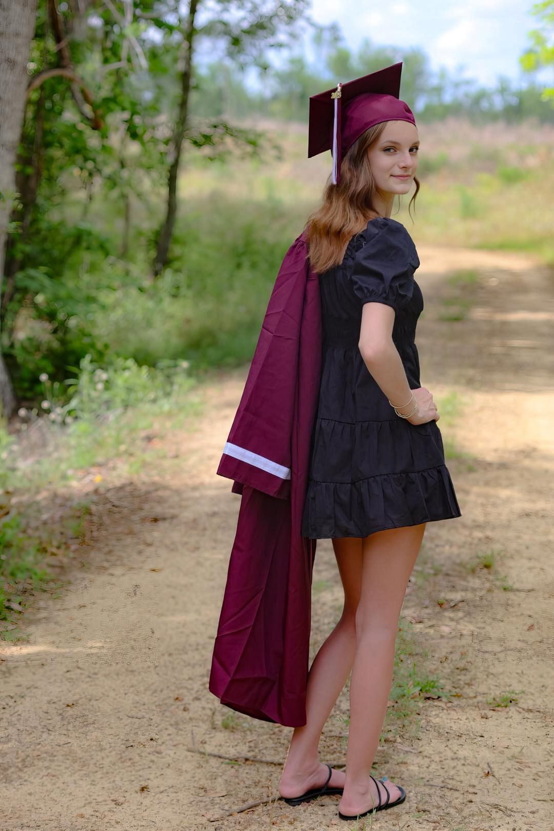 Smiling young woman in maroon graduation cap carrying gown on wooded dirt path.