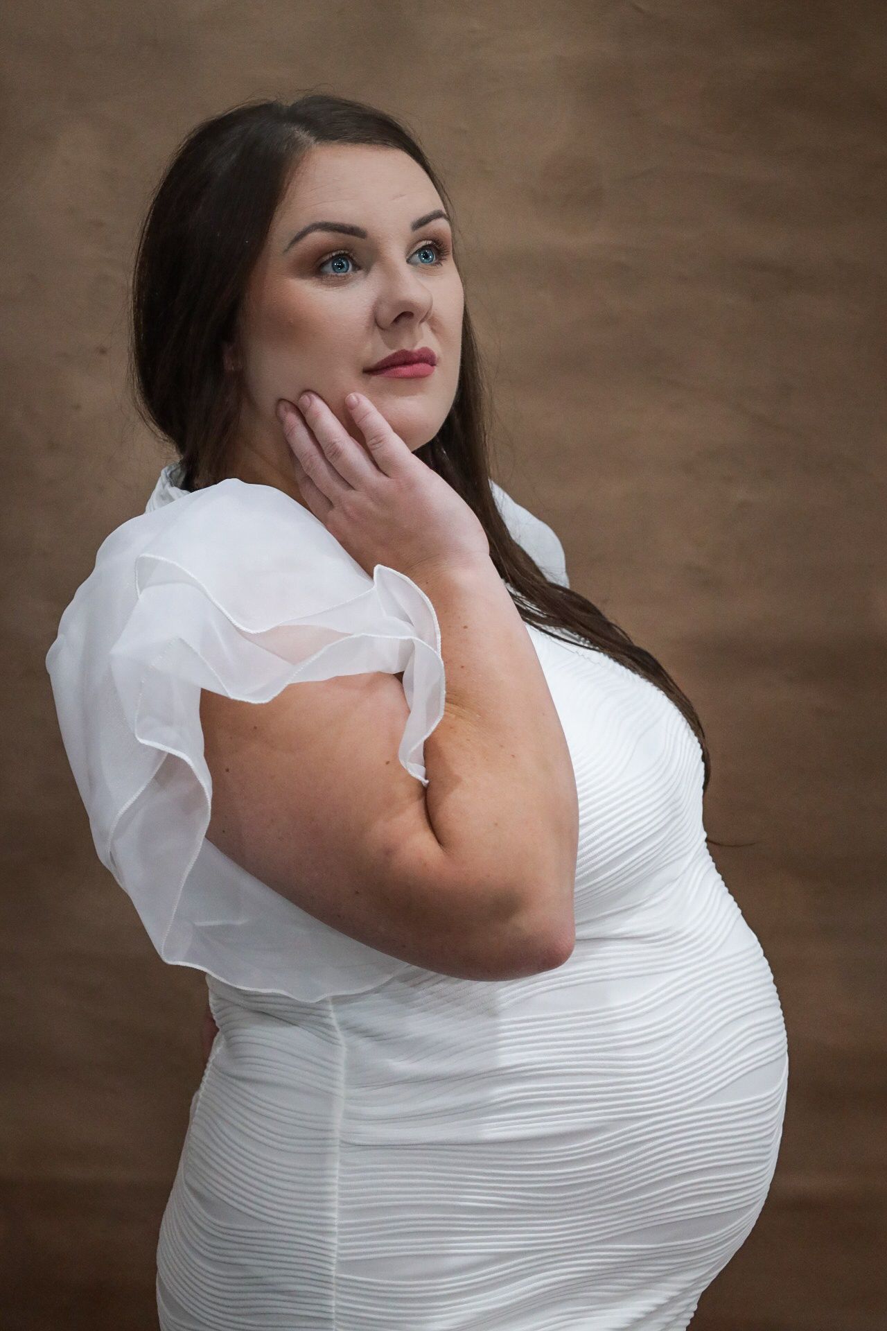 Plus-size woman in white ruffled dress posing thoughtfully with hand on chin against brown background.