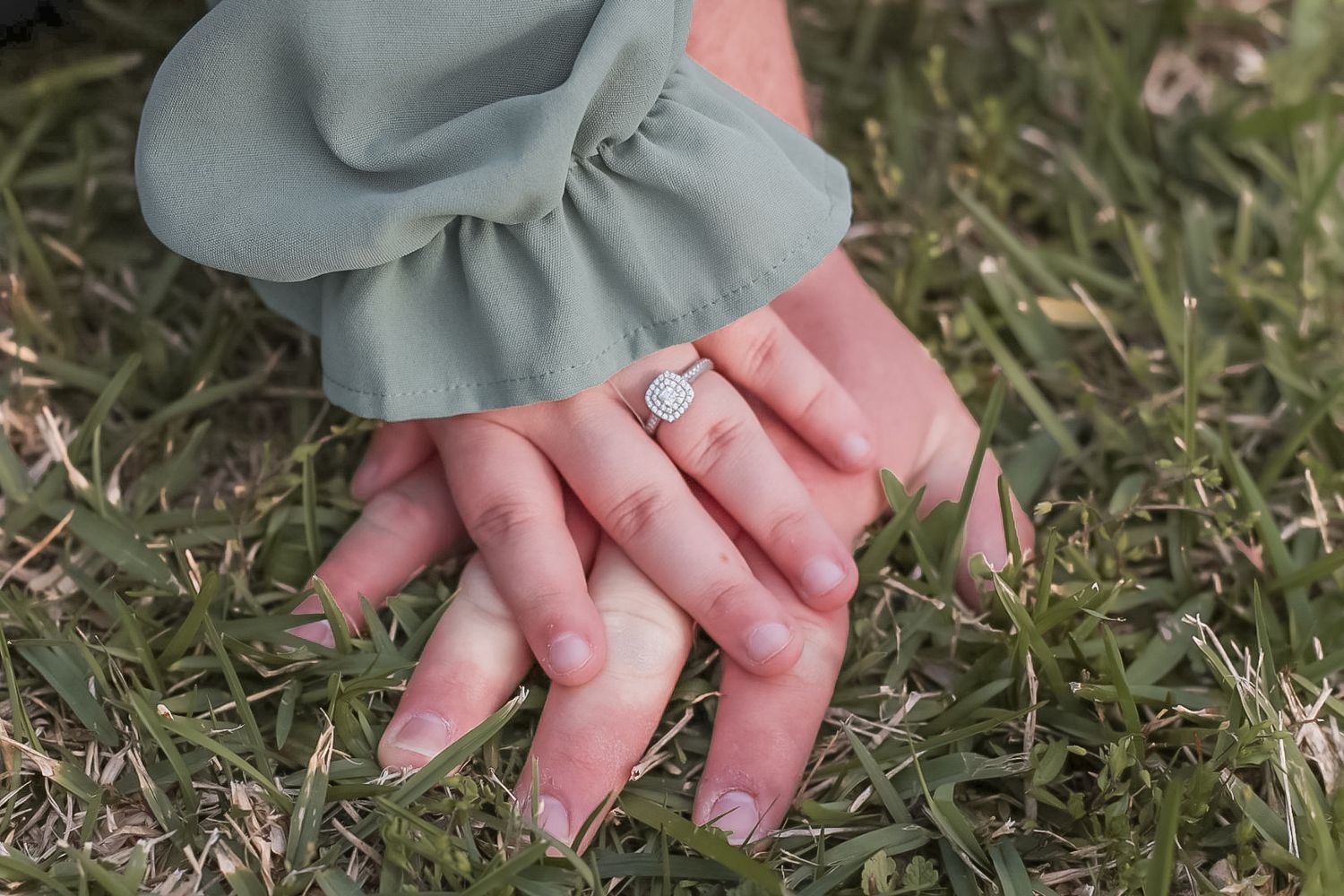 Two hands resting on green grass, one wearing a sparkling diamond engagement ring, with a sage green ruffled sleeve visible.