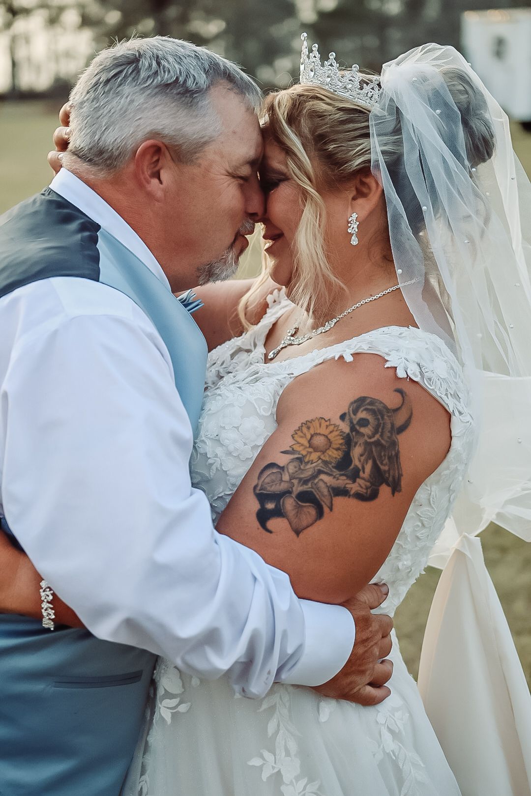 Tender wedding moment showing bride in white lace dress with visible floral arm tattoo and veil sharing embrace with partner.