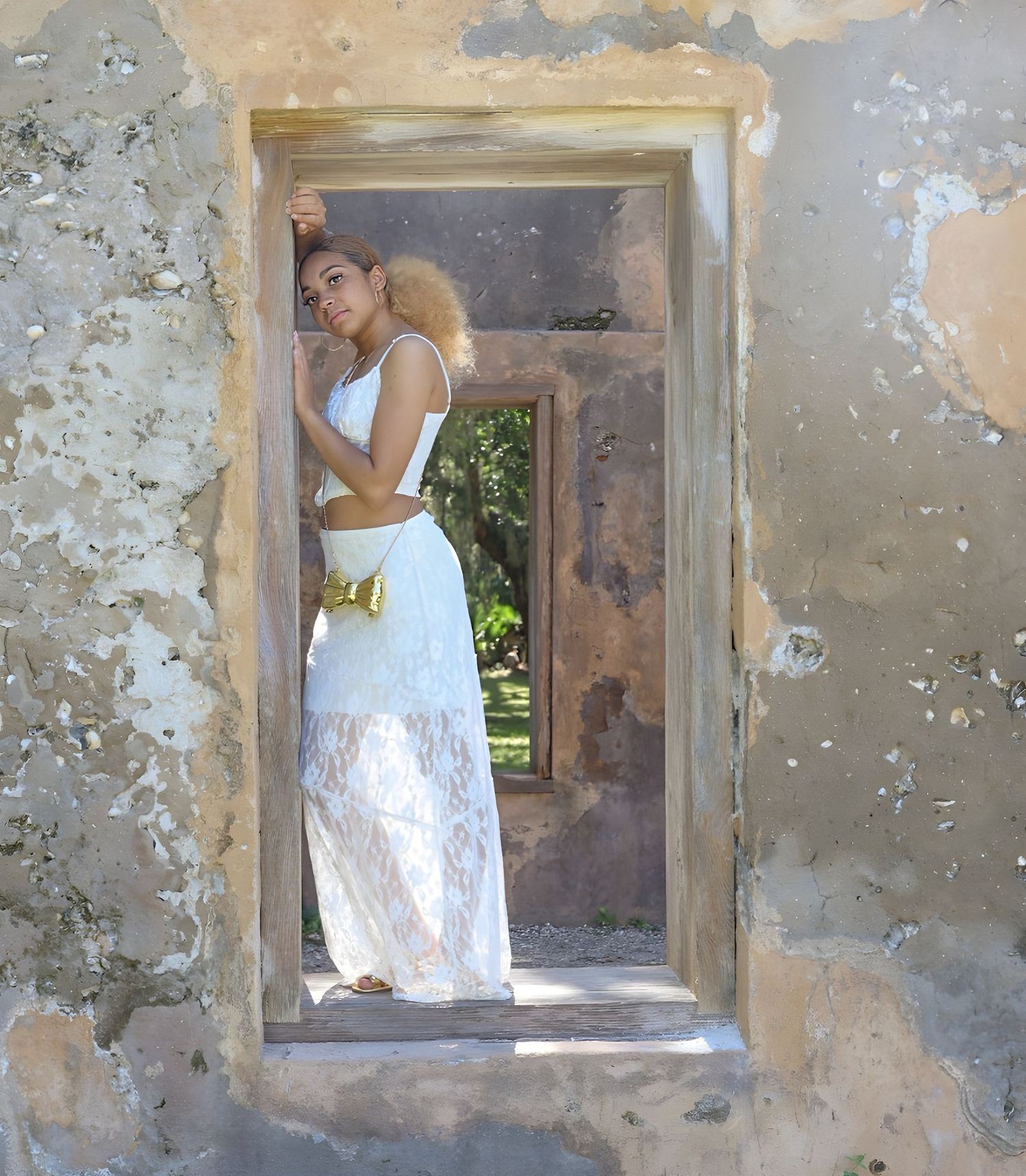Figure in a white dress poses in the stone doorway of a weathered building with peeling walls and greenery visible beyond.