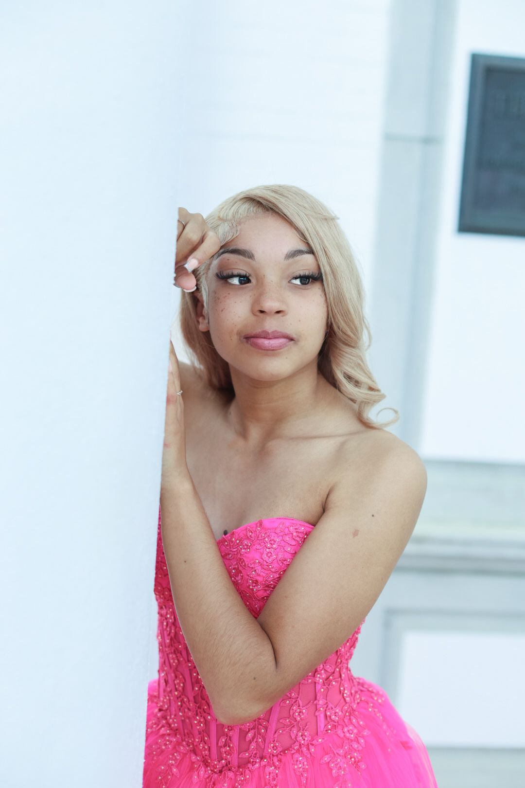 Woman in bright pink strapless dress poses indoors against white wall.