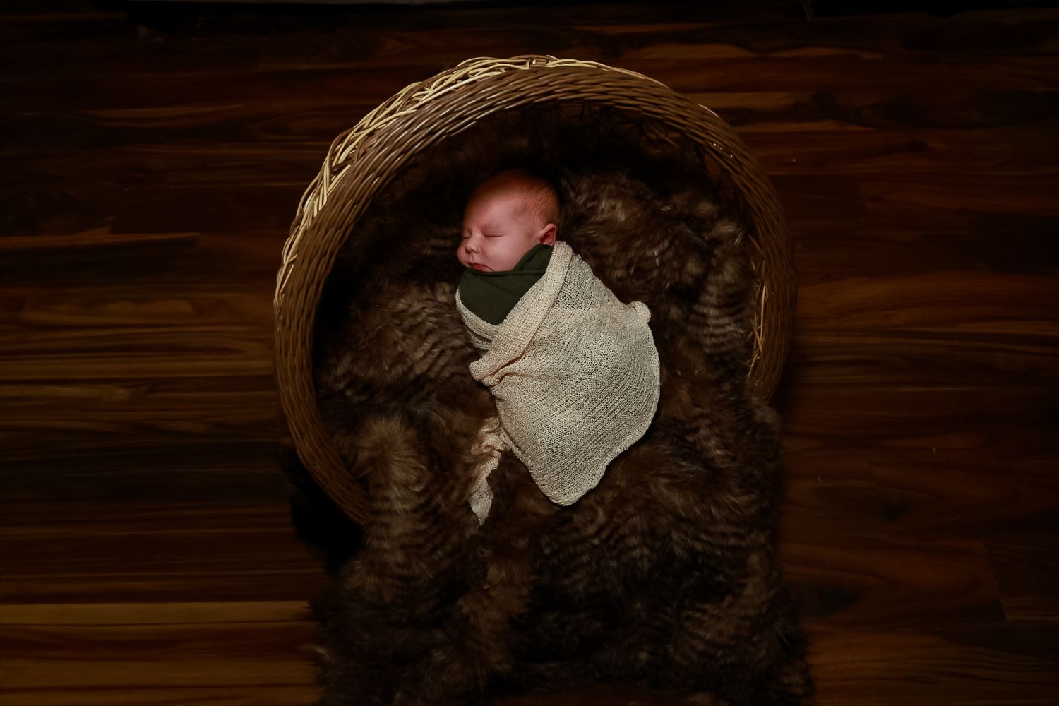 A newborn baby sleeps peacefully in a round wooden basket lined with brown fur and white fabric.