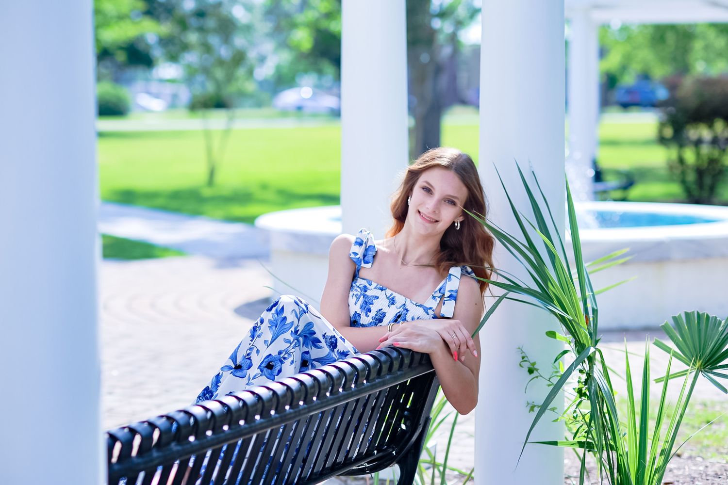 Smiling young woman in blue floral dress seated on bench near white columns.