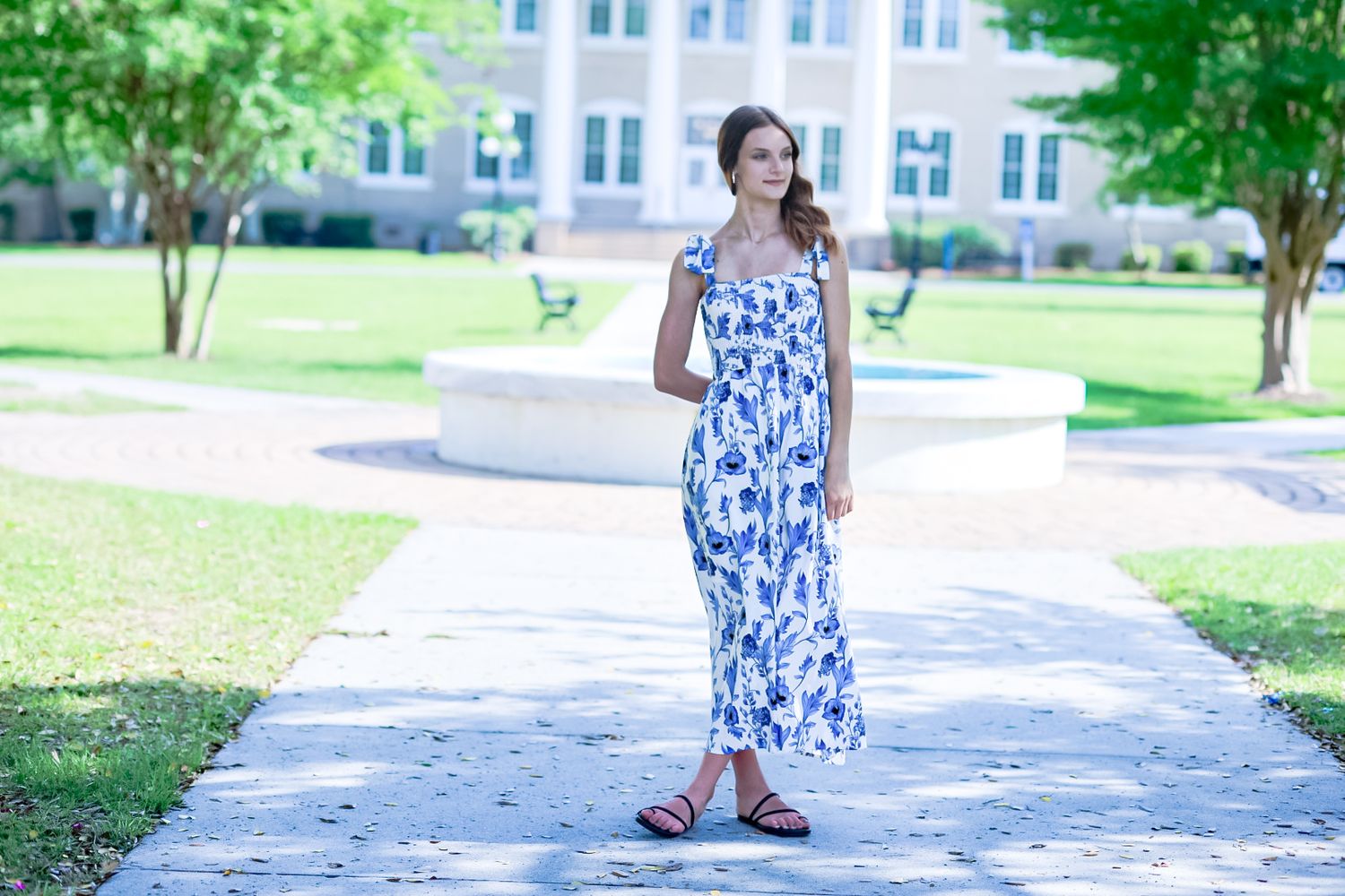 Young woman in floral summer dress walks down a tree-lined campus pathway.