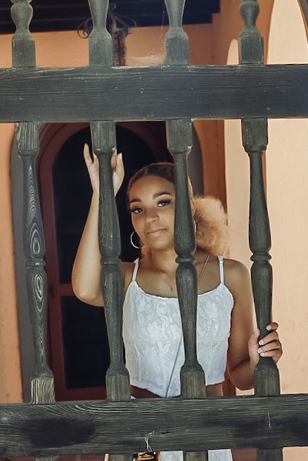 A person in a white top poses between wooden railings on a porch or balcony.