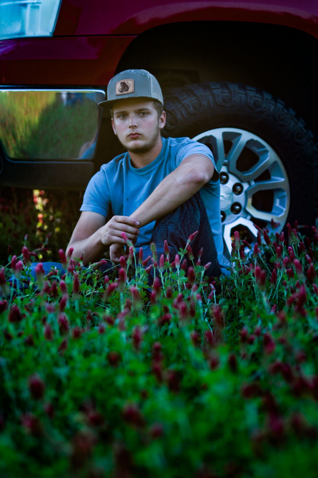 Someone wearing a baseball cap sits in a field of red clover flowers next to a truck tire at dusk.
