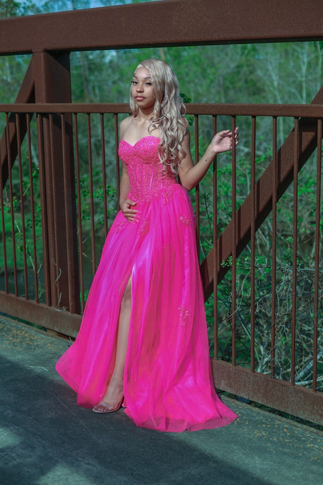 Woman in flowing hot pink formal gown poses on wooden bridge deck surrounded by greenery.