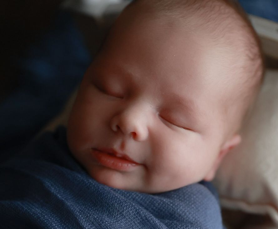 A sleeping newborn baby with rosy cheeks rests peacefully against a blue fabric background.