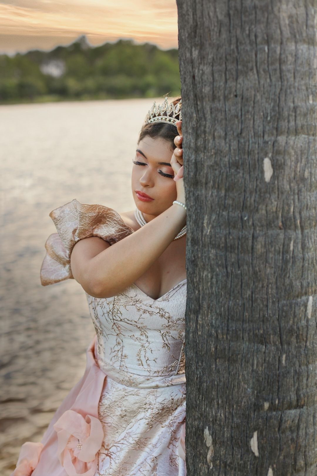 Young woman in elegant white and gold gown with tiara leans against palm tree on beach at sunset.