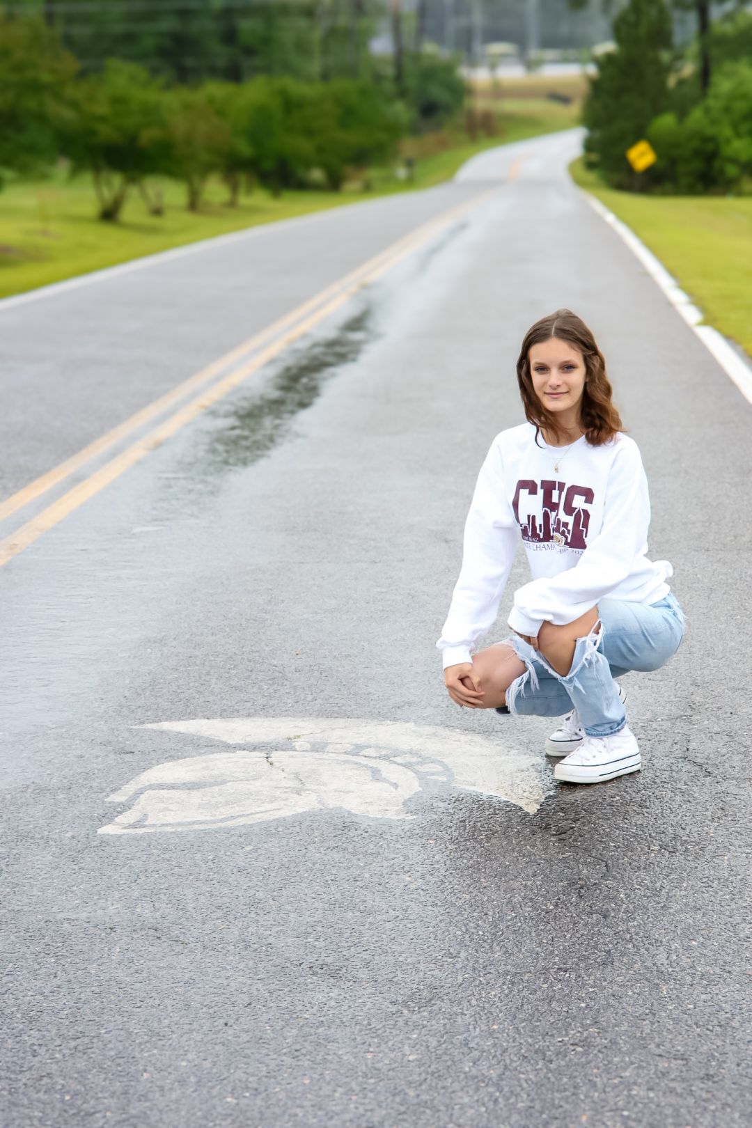 Teen girl in white CHS sweatshirt and ripped jeans crouching on wet road for senior portrait photo.