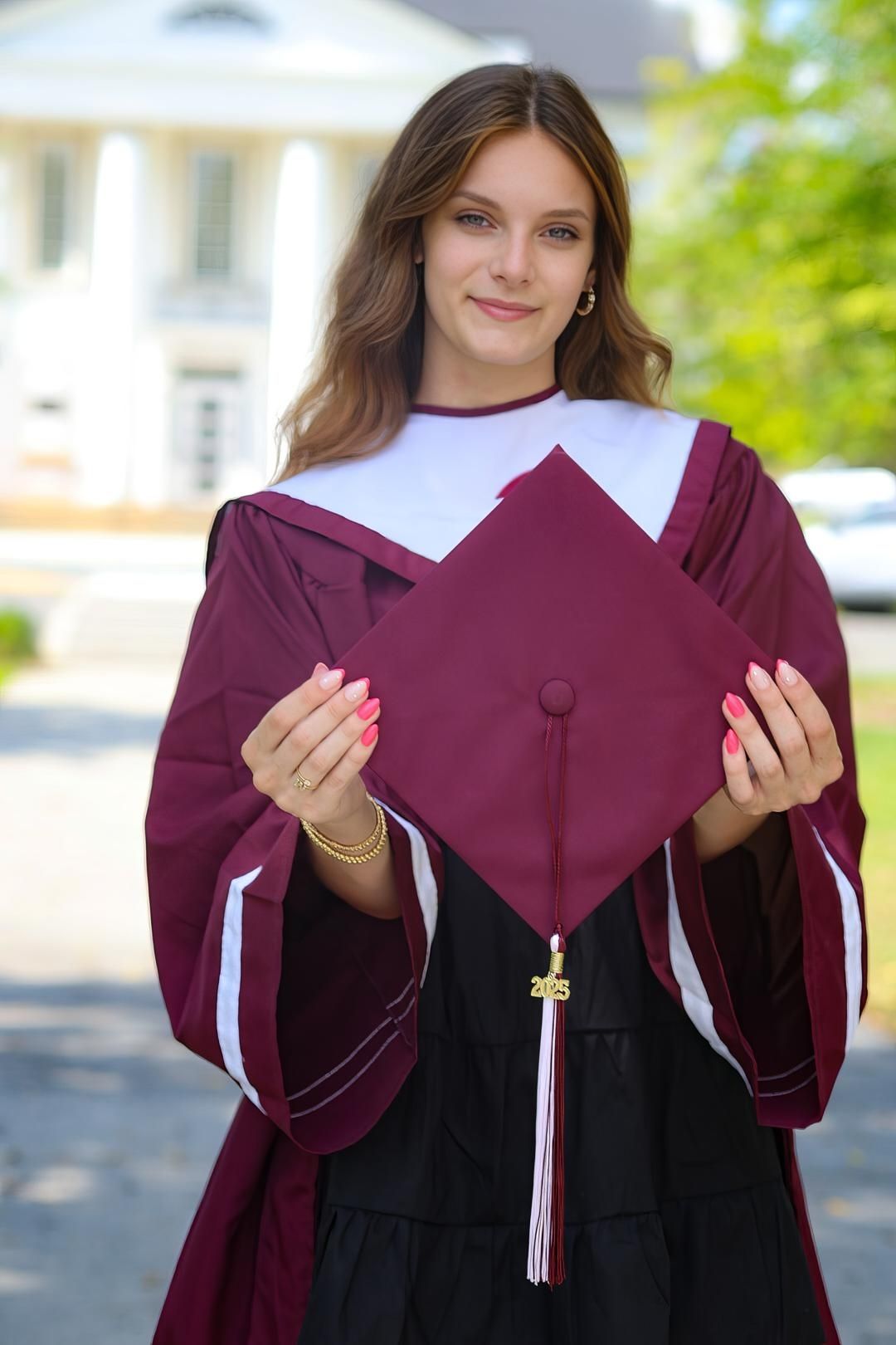 Young woman in maroon graduation cap and gown holding mortarboard toward camera outdoors.