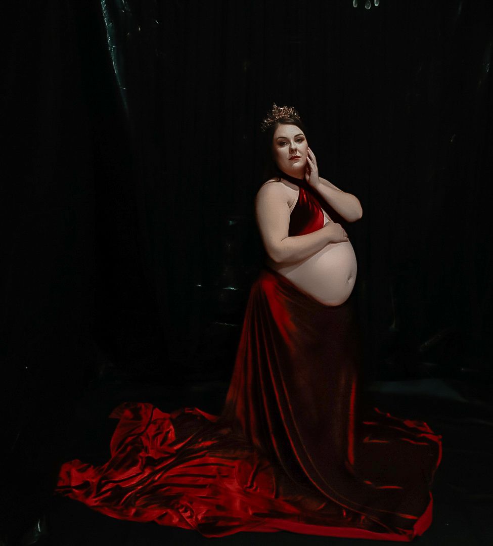 Pregnant woman in red velvet gown with crown poses elegantly against dark backdrop in maternity photo.