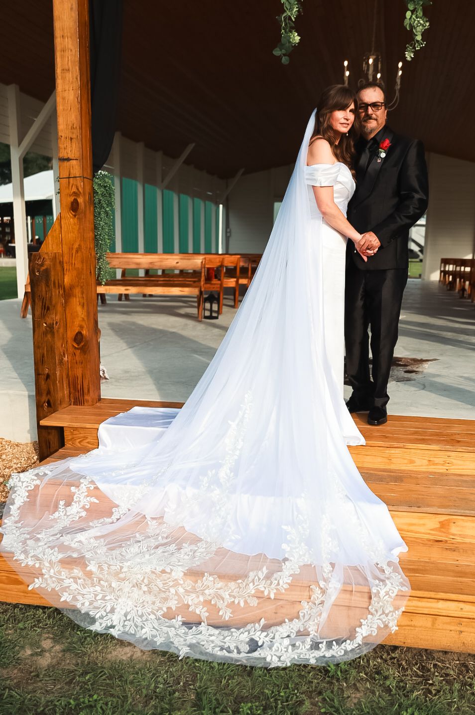Bride in white off-shoulder gown with long lace veil stands with groom in black suit at outdoor wedding venue.