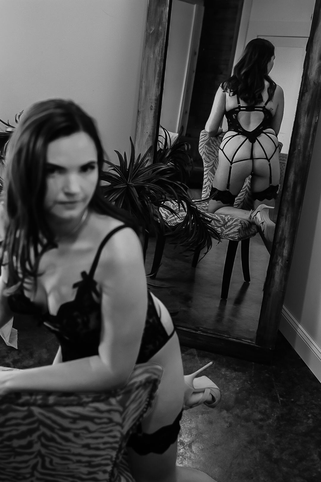 Black and white boudoir photo of a woman in lingerie seated near a mirror reflecting her back view.