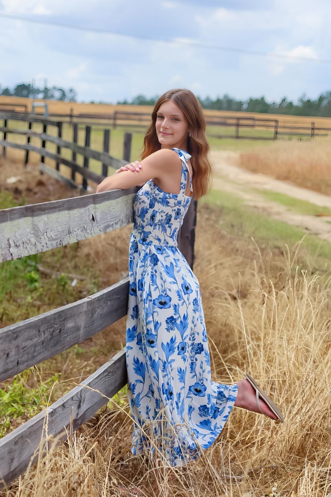 A floral blue and white maxi dress photographed against a rustic wooden fence in a countryside setting.