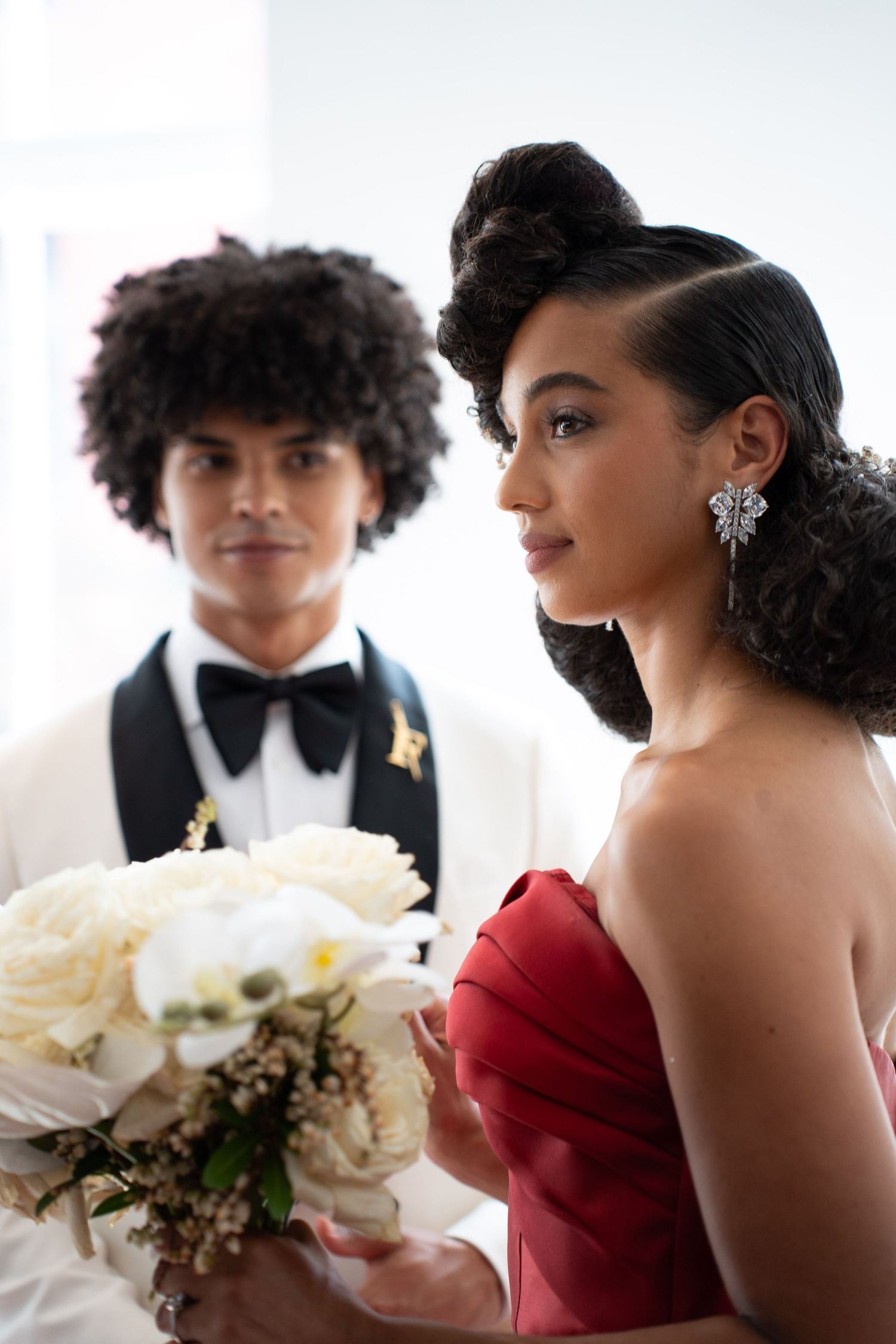 Close-up of woman in red strapless gown with butterfly earring holding white bouquet, man in tuxedo blurred behind.