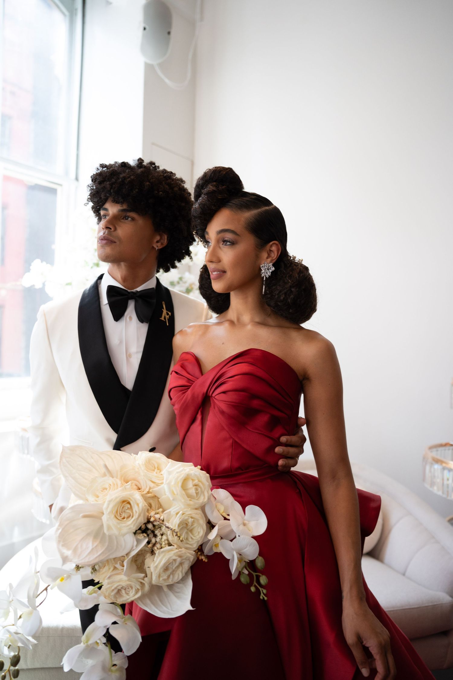 Stylish bride in red strapless gown and groom in white tuxedo holding white floral bouquet.